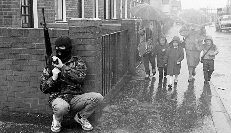 Photo of an armed IRA fighter hiding behind a wall with a young family coming his way