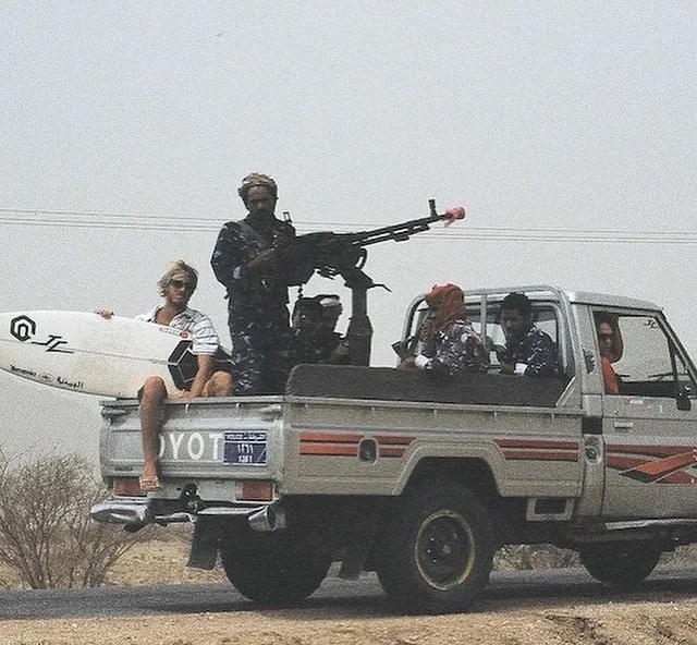 Photo of a surfer between armed fighters on the back of a Yemeni pickup truck