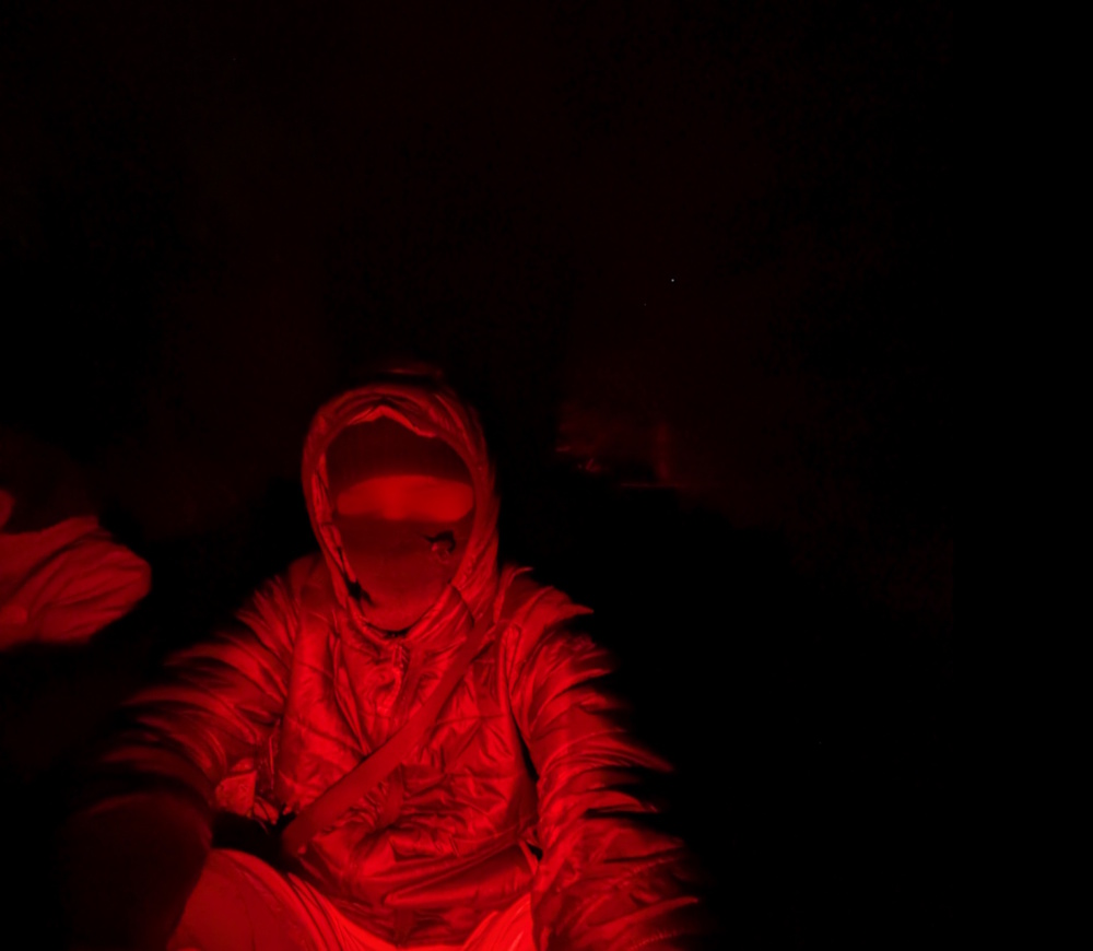 A person wearing dark clothes and a balaclava sitting and looking at the camera in dim red light